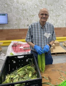 Picture of volunteer, Jim, standing behind a table with gloves on while holding two ears of corn.